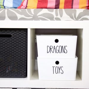 bench with cushion and colorful pillows. under bench two white bins with black labels that say dragons and toys to demonstrate toy storage of action figures