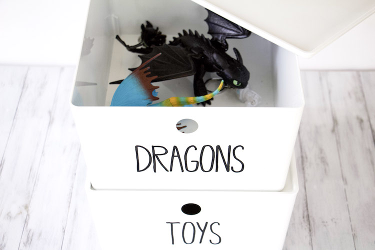 Two white boxes stacked on top of each other to demonstrate toy storage of action figures. Top bin is open to show dragons that coinsides with label on bin